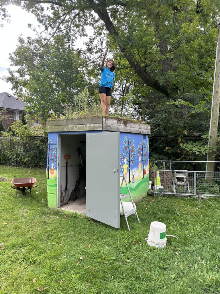 Teenage girl stands on shed green roof with gardening tool in hand. 