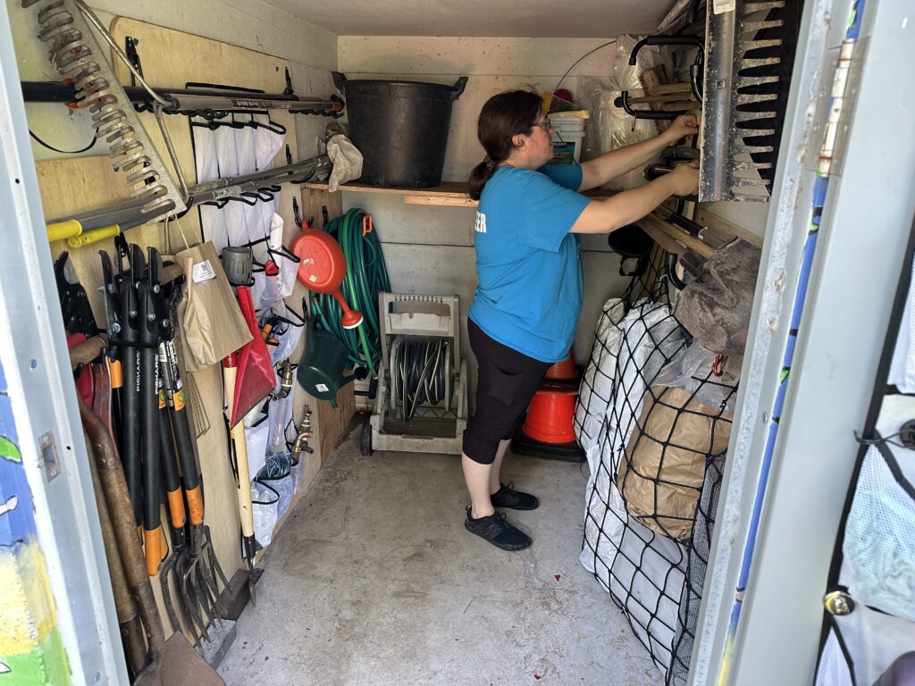 Sara in the shed with the walls lined with tools hanging on hooks. 